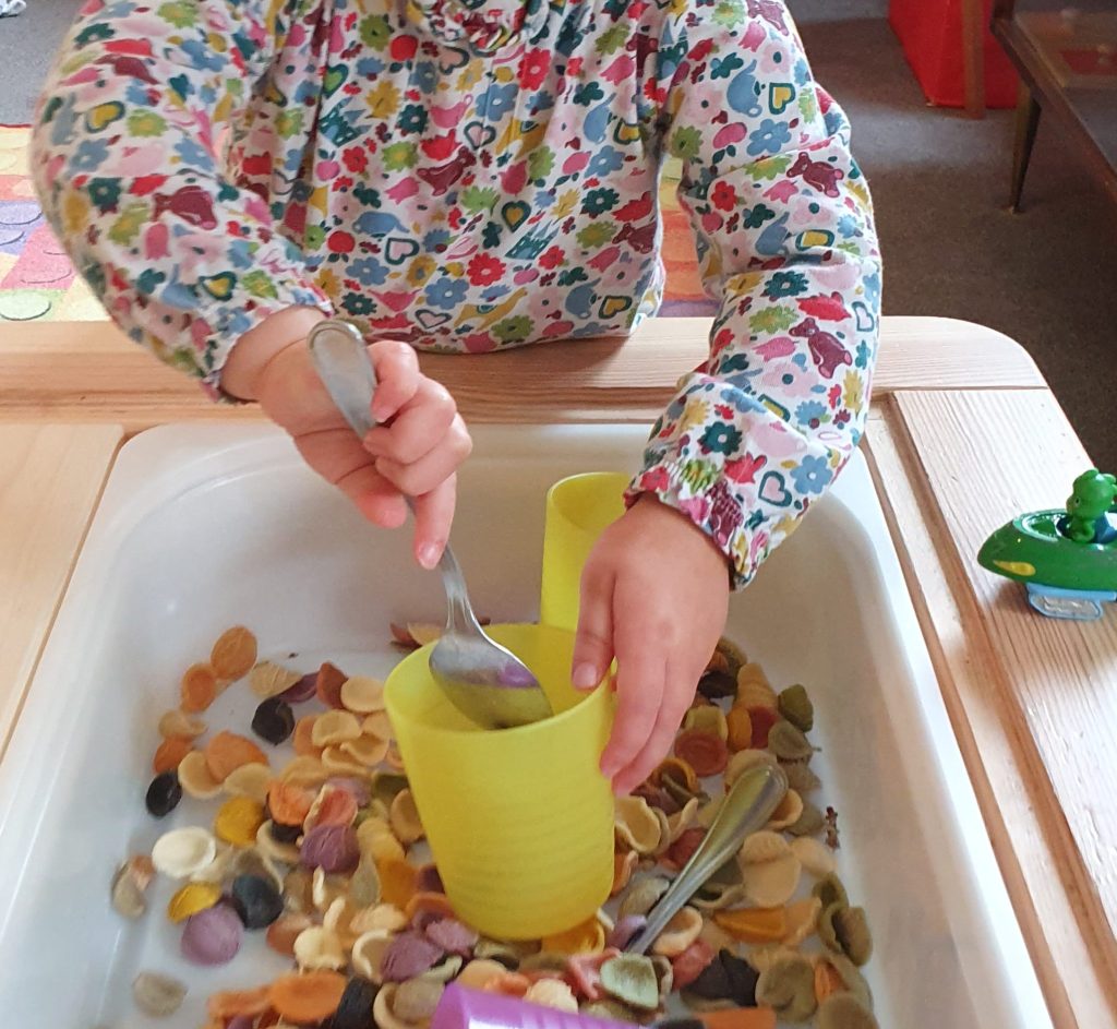 Child scooping dry coloured pasta with a spoon into a yellow plastic cup