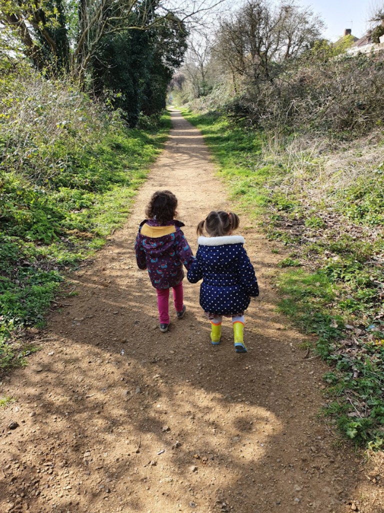 Two children holding hands, walking along a path between shrubs and trees